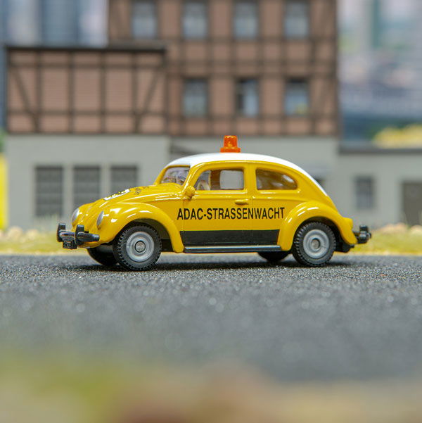 VW Käfer ADAC in gelb mit Print  ADAC Straßenwacht. Steht auf grauem Asphalt  mit Häusern im Hintergrund