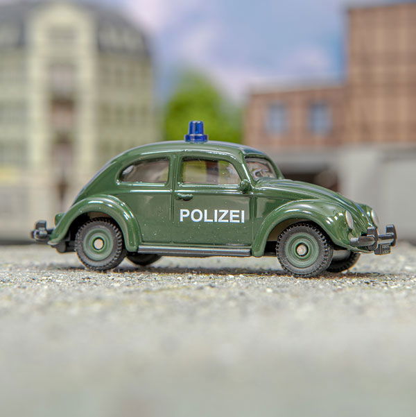 VW Käfer in dunkelgrüner Polizei-Version steht auf Asphalt  vor verschwommenem Häuserhintergrund
