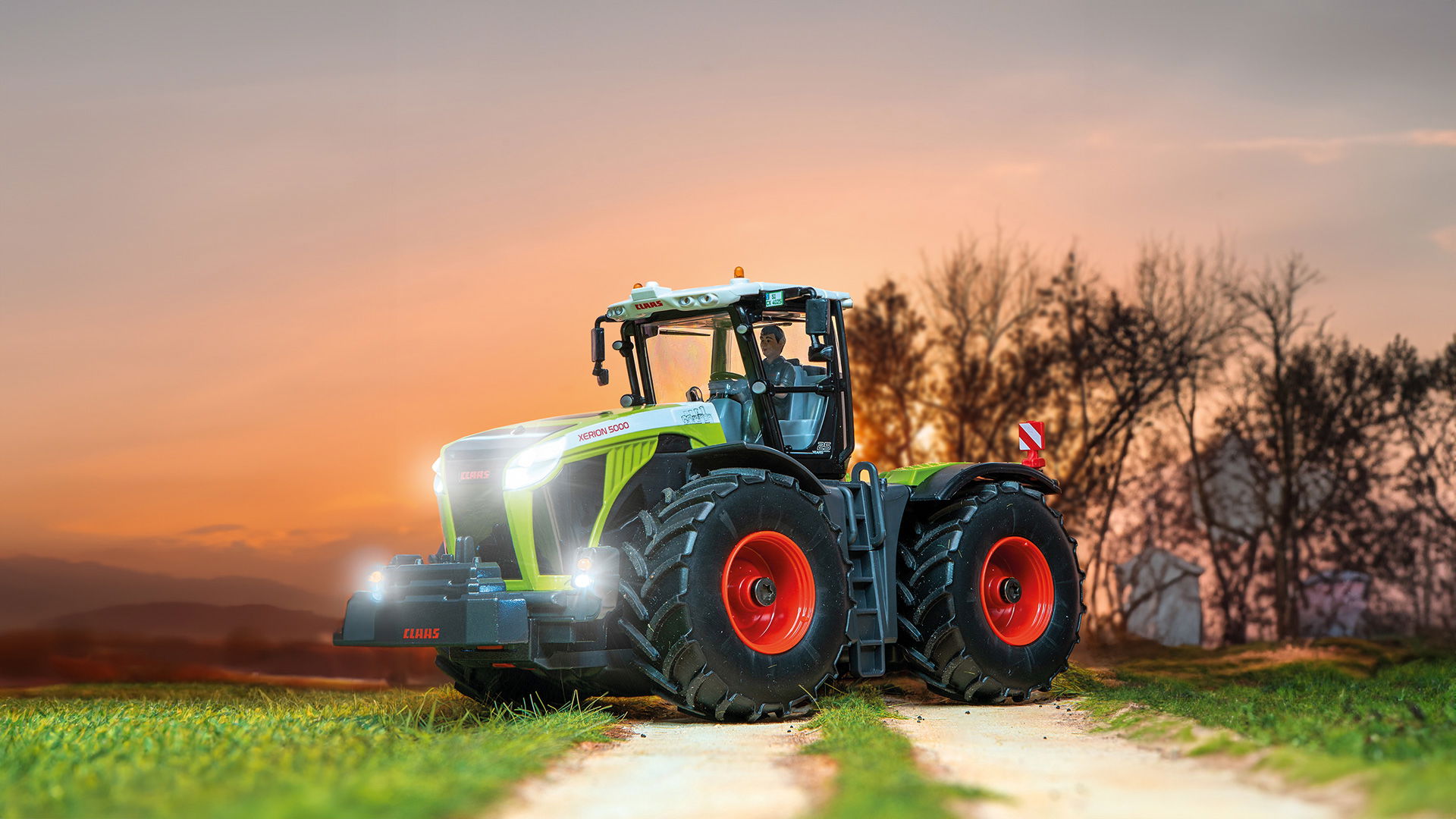 Der SIKUCONTROL Claas Xerion steht auf einem Feldweg mit angeschalteter Beleuchtung. Im Hintergrund ist ein Sonnenuntergang zu sehen