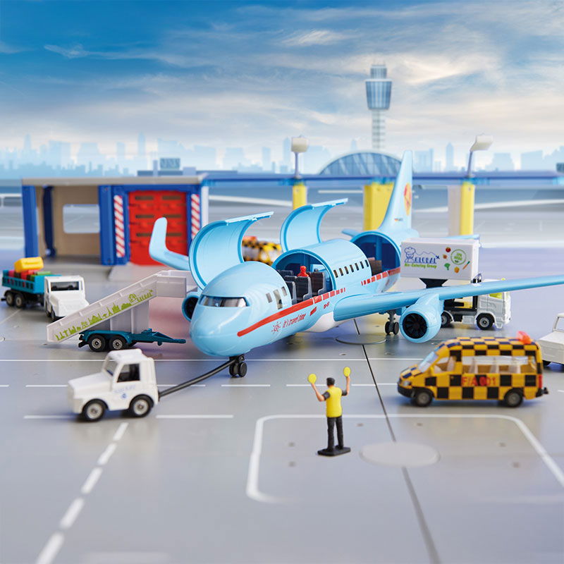 Light blue commercial aircraft stands on a grey ground with its flaps open. Various airport vehicles and an air traffic controller stand around the aircraft. A tower and a skyline can be seen in the background.